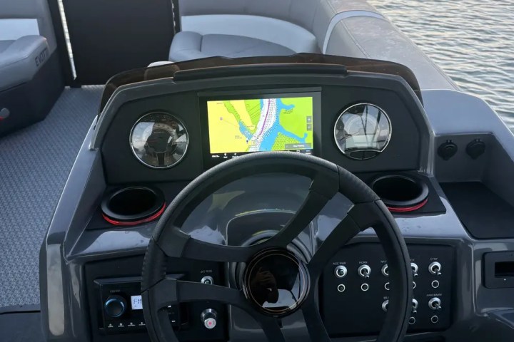 Boat helm with steering wheel and electronic display, sunset over calm water in background.