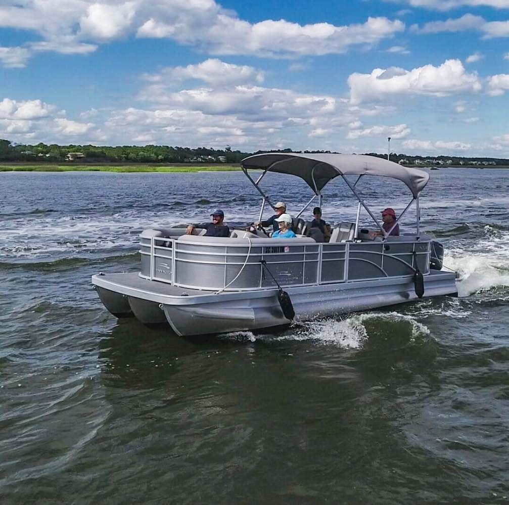 Pontoon boat with canopy on water, four passengers onboard, cloudy sky.